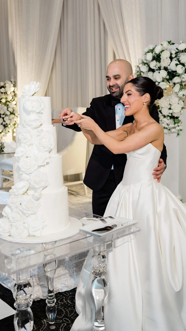 Smiling bride and groom cutting a white tiered wedding cake at their reception.