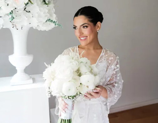 Bride in a lace robe holding a bouquet of white flowers beside a floral arrangement.